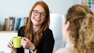 A woman with glasses, holding a lime green mug, is smiling to another woman.